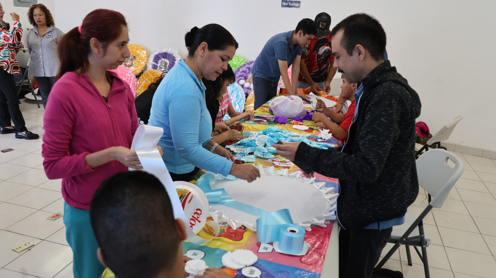Personas elaboran coronas con flores de papel y listones alrededor de una mesa.