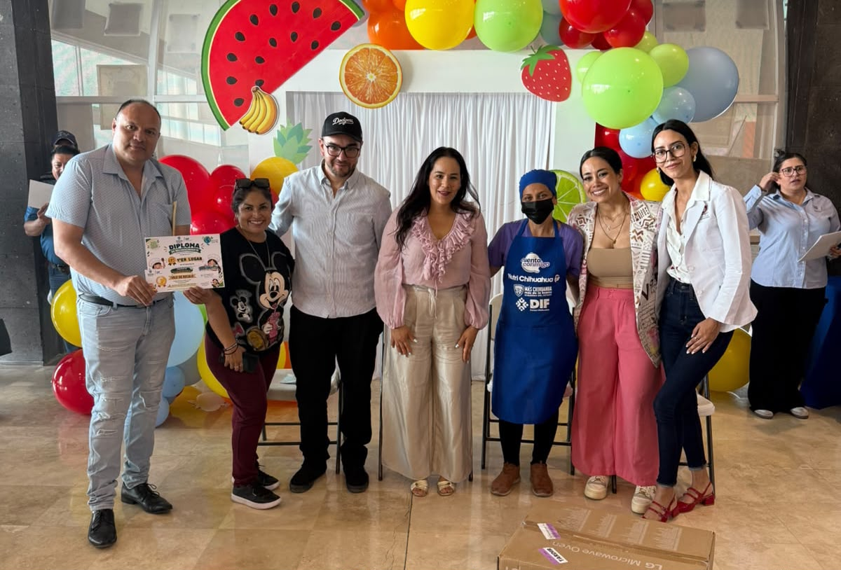 Grupo de personas posa con un diploma frente a una decoración de frutas y globos de colores.